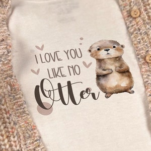 Könnte beinhalten: Weißer Baby-Strampler mit einer braunen Otter-Illustration und dem Text "Ich liebe dich wie keine andere". Der Strampler wird mit einem beigefarbenen Cardigan kombiniert.