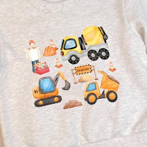 Op de afbeelding: Een grijs sweatshirt met een bouwthema. Het ontwerp bevat een gele betonmixer, een gele kiepwagen, een bouwvakker en een bord met de tekst "Work in Progress".