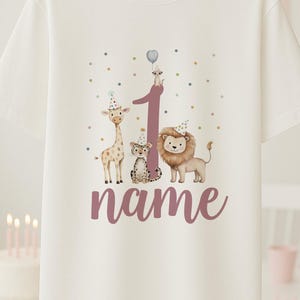 May include: White t-shirt with a birthday theme. The shirt features a large number 1 with a giraffe, leopard, and lion wearing party hats. The word "name" is below the number. Colorful polka dots and a meerkat with a balloon complete the design.