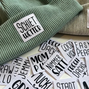 Könnte beinhalten: Eine Sammlung weißer Stoffpatches mit schwarzem Text. Die Patches zeigen verschiedene Sätze, darunter "Mom", "Satt", "Stadt Kind", "Frech Dachs", "Schielt Wetter" und "Girl Power".