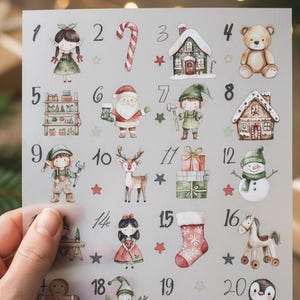 Könnte beinhalten: Ein transparentes Blatt mit Aquarellillustrationen für einen Weihnachtskalender. Das Blatt zeigt nummerierte Bilder von festlichen Figuren und Objekten, darunter der Weihnachtsmann, Elfen, ein Schneemann und ein Rentier. Das Blatt wird von einer Hand gehalten.