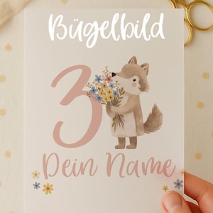 Pode incluir: Um cartão de felicitações com o texto "Bügelbild" e "3" em rosa. Apresenta uma raposa de desenho animado segurando flores. O texto "Dein Name" também está presente. Tesoura dourada e fita no fundo.