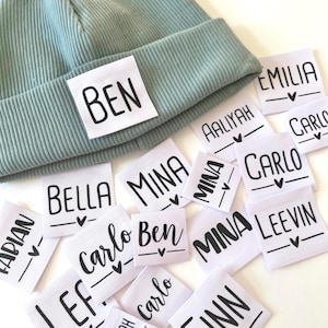 May include: A collection of white fabric labels with black text and a small heart design. The labels feature various names, including Ben, Bella, Mina, Carlo, Aaliyah, Emilia, and Leevin. One label is attached to a light blue beanie.