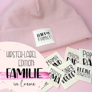May include: A set of iron-on labels with the words "Happy Family", "Mama", "Papa", "Mädchen", "Kleiner Bruder", "Kleine Schwester" and "Mädchen" in black text on a white background. The labels are on a pink surface.