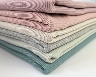 Puede incluir: Una pila de cinco tejidos de punto de canalé de diferentes colores. Los tejidos están doblados cuidadosamente y son en tonos de rosa, crema, gris y azul.