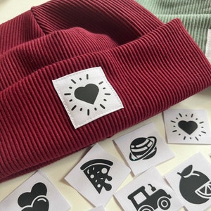 Puede incluir: Un gorro burdeos con un parche blanco que presenta un corazón negro y un diseño de rayos de sol. El gorro está rodeado de varios parches blancos con diseños negros, incluyendo un corazón, un planeta, una rebanada de pizza, un tractor, una fruta y un sol con un corazón.