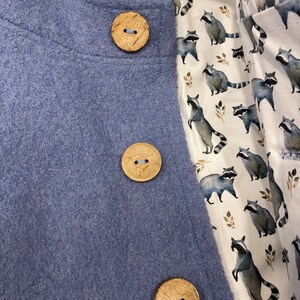 May include: A blue fleece jacket with three wooden buttons. The jacket is lined with a white fabric featuring a repeating pattern of raccoons.