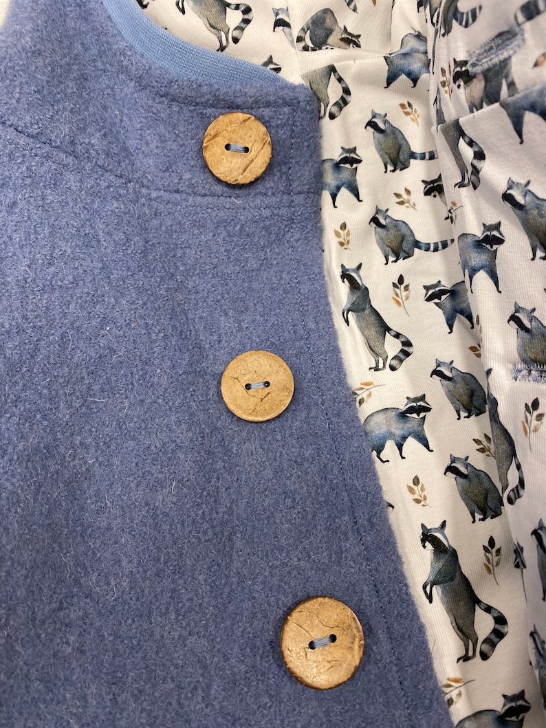 May include: A blue fleece jacket with three wooden buttons. The jacket is lined with a white fabric featuring a repeating pattern of raccoons.