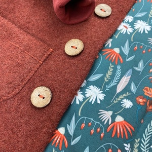 May include: A close-up of a rust-coloured fabric with three wooden buttons. The fabric is folded over a teal fabric with a floral pattern.