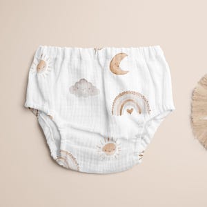 Op de afbeelding: Witte babybloomers met een herhalend patroon van aquarel zonnen, regenbogen, wolken en manen. De bloomers zijn gemaakt van een zachte, getextureerde stof. Het ontwerp heeft een neutraal kleurenpalet.