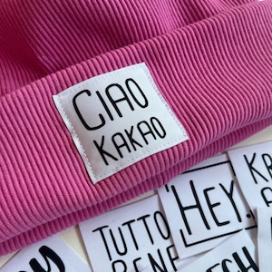 Könnte beinhalten: Eine rosafarbene Strickmütze mit einem weißen Patch, auf dem in schwarzen Buchstaben "CIAO KAKAO" steht.