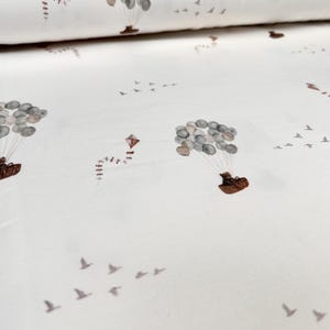 Pode incluir: Tecido branco com um padrão repetido de balões de ar quente feitos de papel castanho e balões cinzentos e brancos. Os balões estão amarrados com cordas e têm uma pequena cesta castanha por baixo. Também há pequenos pássaros castanhos a voar no padrão.