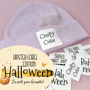 Könnte beinhalten: Eine lila gerippte Strickmütze mit einem weißen Etikett mit der Aufschrift "Creepy Cutie". Zusätzliche Etiketten mit Text sind zu sehen. Das Bild enthält auch den Text "Hipster-Label Edition: Halloween" und "Zu süß zum Gruseln!" mit einer Kürbisgrafik.