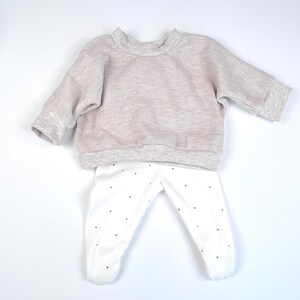 May include: A two-piece baby outfit in grey and white. The top is a waffle knit long-sleeved sweater. The bottom is white leggings with small red polka dots.
