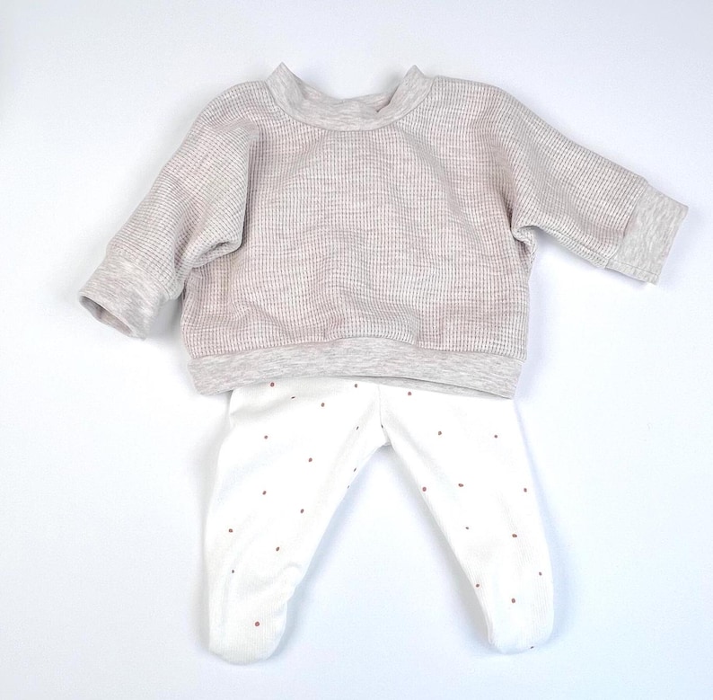 May include: A two-piece baby outfit in grey and white. The top is a waffle knit long-sleeved sweater. The bottom is white leggings with small red polka dots.