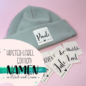Könnte beinhalten: Eine hellgraue Mütze mit einem weißen Etikett, auf dem "Pauli" mit einem kleinen Herz steht. Die Mütze ist umgeben von weißen Stoff-Etiketten mit Namen in schwarzer Tinte, darunter "JUVENT", "due Matilde", "Jule" und "Karl". Der Text "HIPSTER-LABEL EDITION: NAMEN in Weiß und Creme" ist in der linken unteren Ecke des Bildes sichtbar.