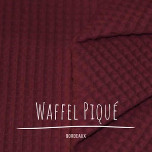 May include: Close-up of a burgundy waffle pique fabric. The fabric has a textured, grid-like pattern. The text "WAFFEL PIQUÉ" is displayed in white, with "BORDEAUX" underneath.