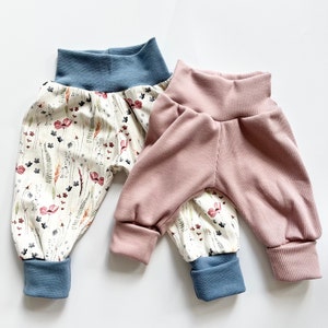 May include: Two pairs of pants. One pair features a cream background with a multi-colored floral and botanical print, finished with solid blue ribbed cuffs and waistband. The second pair is solid dusty pink ribbed fabric with blue cuffs.