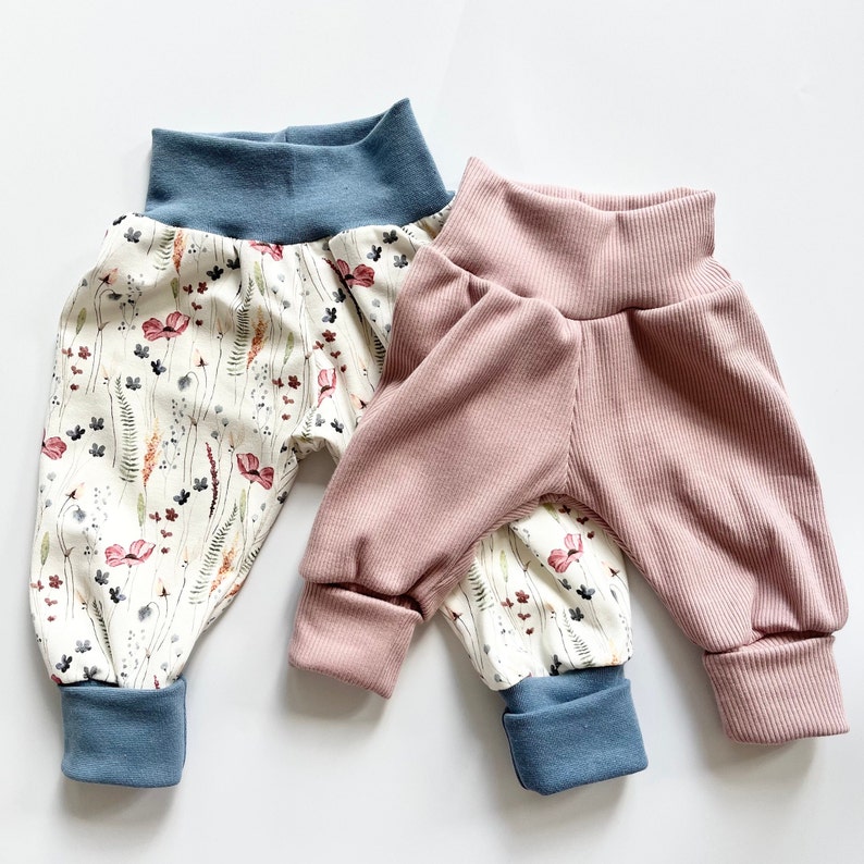 May include: Two pairs of pants. One pair features a cream background with a multi-colored floral and botanical print, finished with solid blue ribbed cuffs and waistband. The second pair is solid dusty pink ribbed fabric with blue cuffs.