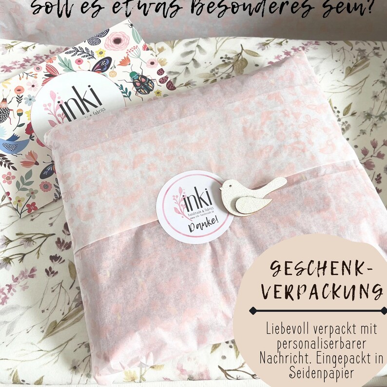 May include: A rectangular package wrapped in light pink tissue paper, secured with a round white sticker featuring the text inki and Danke, and a small wooden bird cutout. The package rests on floral fabric with the text Soll es etwas Besonderes sein?
