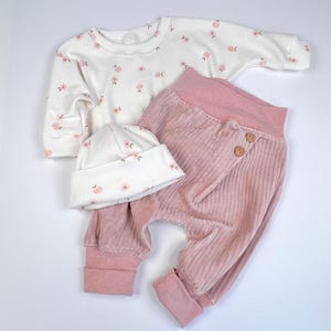 May include: A baby outfit set with a white long-sleeved shirt with a peach floral print, pink corduroy pants, and a white beanie with a peach floral print.