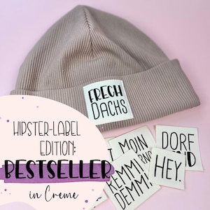 Könnte beinhalten: Eine beige Mütze mit einem weißen Etikett, auf dem "FRECH DACHS" steht. Die Mütze ist umgeben von weißen Stoff-Etiketten mit schwarzem Text, darunter "DORF", "HEY", "REMM", "DEMM", "MOIN". Die Etiketten befinden sich auf einem rosa Hintergrund.