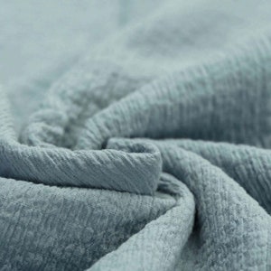 Può includere: Primo piano di un tessuto morbido e testurizzato di colore azzurro chiaro. Il tessuto ha un aspetto stropicciato e raggrinzito.