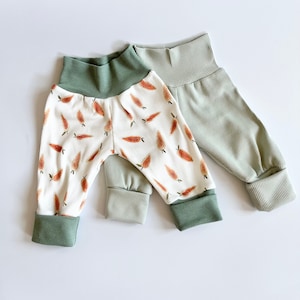 May include: Two pairs of ribbed pants. One pair is white with an orange carrot print, featuring a solid sage green waistband and matching ankle cuffs. The second pair is solid sage green with a ribbed texture, a high waistband, and folded cuffs.