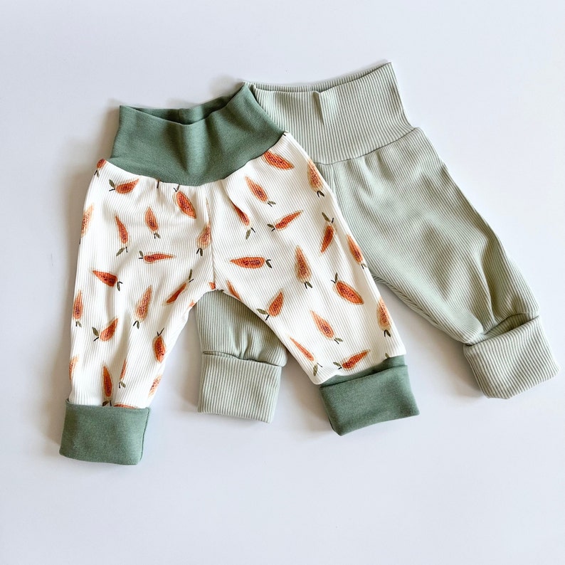 May include: Two pairs of ribbed pants. One pair is white with an orange carrot print, featuring a solid sage green waistband and matching ankle cuffs. The second pair is solid sage green with a ribbed texture, a high waistband, and folded cuffs.