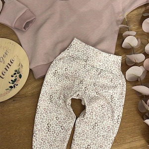 May include: A pink and white striped long-sleeved shirt with a matching pair of leggings featuring a pink and white leopard print. A pink headband with a leopard print is also included. The text "Den Mama" is visible on a wooden sign.