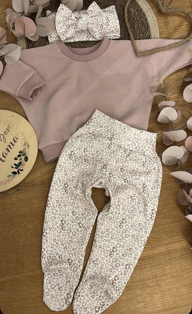 May include: A pink and white striped long-sleeved shirt with a matching pair of leggings featuring a pink and white leopard print. A pink headband with a leopard print is also included. The text "Den Mama" is visible on a wooden sign.