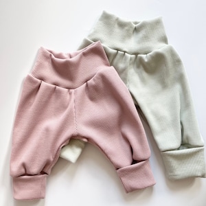 May include: Two pairs of ribbed pants, one in a soft dusty pink and the other in a pale sage green. Both pairs feature a high, wide waistband and cuffed ankles, constructed from a textured, vertically ribbed fabric with a relaxed fit.