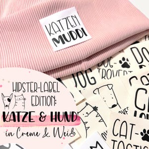 Könnte beinhalten: Eine rosa Strickmütze mit einem weißen Etikett, auf dem "KATZEN MUDDI" steht. Die Mütze ist von verschiedenen Stoff-Etiketten mit schwarzem Text und Illustrationen von Katzen und Hunden umgeben. Die Etiketten sagen "HIPSTER-LABEL EDITION: KATZE & HUND in Creme & Weis", "JOG", "DOG-TROVER", "CAT-TACTIC" und "DOG".