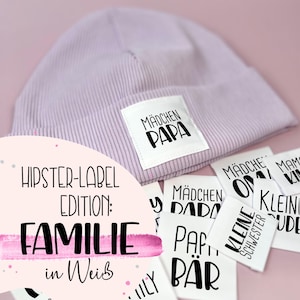 May include: A set of white iron-on labels with black text. The labels feature the words "Mädchen Papa", "Mädchen Oma", "Mädchen Dada", "Papa Bär", "Kleine Schwester", "Kleine Bruder", and "Mädchen" in a variety of fonts. The labels are arranged on a pink surface.
