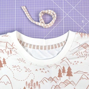 May include: A white t-shirt with a brown mountain and forest print. The neckline is finished with a double-fold beige and white striped trim.