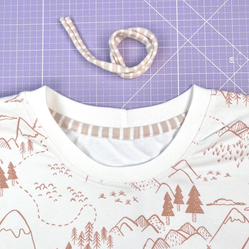 May include: A white t-shirt with a brown mountain and forest print. The neckline is finished with a double-fold beige and white striped trim.