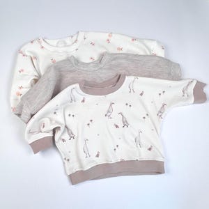 May include: Three long-sleeved shirts for babies. The top shirt is white with pink flowers, the middle shirt is grey with a waffle texture, and the bottom shirt is white with a pink trim and a pattern of brown ducks.