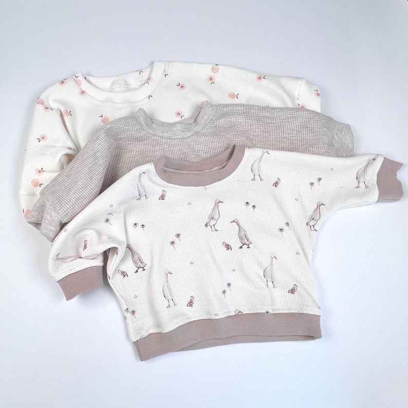 May include: Three long-sleeved shirts for babies. The top shirt is white with pink flowers, the middle shirt is grey with a waffle texture, and the bottom shirt is white with a pink trim and a pattern of brown ducks.
