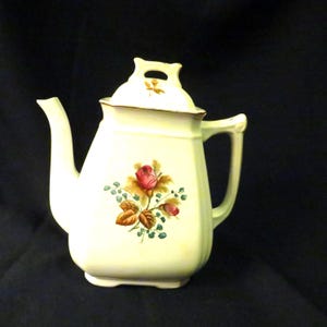 May include: Cream-colored ceramic teapot with a lid and handle. The teapot features a floral design with a red rose and green leaves. The spout and handle are curved. The lid has a small opening and a gold trim.