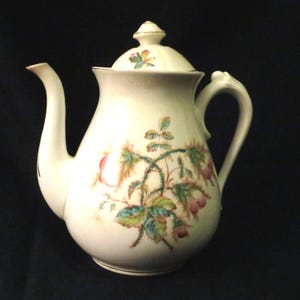 May include: Cream-coloured ceramic teapot with a lid and curved spout. The teapot is decorated with a floral pattern featuring pink, green, and brown botanical elements. The handle has a unique, curved design. A small gold accent is on the lid.