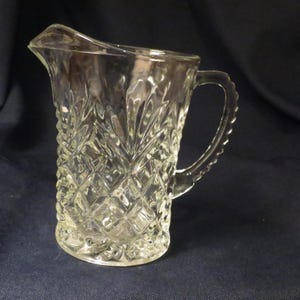 May include: A clear glass pitcher with a diamond pattern. The pitcher has a handle and a wide mouth.