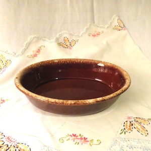 May include: A brown oval baking dish with a glossy finish. The rim has a lighter, speckled color. The dish is empty and sits on a white cloth with floral embroidery.