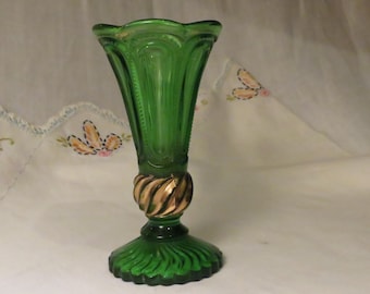 May include: A green glass vase featuring a flared, scalloped rim and vertical ribbed detailing on the body. The stem is accented with a metallic gold-toned twisted band, and the circular base displays a radiating, fan-like pattern in green glass.