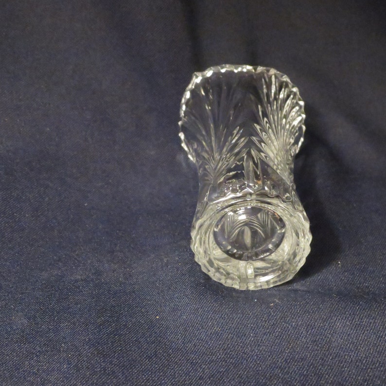 May include: A clear glass bud vase with a cut glass design. The vase has a scalloped rim and a round base.