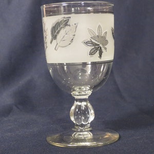 May include: A clear glass goblet with a frosted band and silver leaf design. The goblet has a round base and a stem with a bulbous knob.