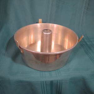 Puede incluir: Un molde para bizcocho bundt redondo de aluminio vintage con un tubo central y dos asas laterales. El molde tiene un acabado metálico brillante y está diseñado para hornear pasteles. El molde está sobre una superficie verde.