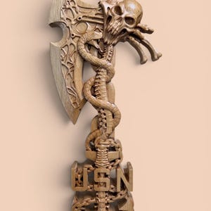 May include: A light brown wooden wall carving featuring a detailed axe head, a skull, and a coiled snake. The handle incorporates a spinal column design, a ship anchor, and the letters USN carved in a block font at the base of the handle.