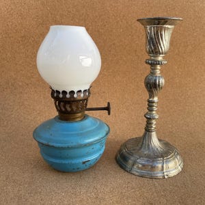 May include: A small oil lamp with a bright blue metal base, a brass burner, and a white glass globe, positioned next to a tall, slender silver-toned metal candlestick holder featuring a spiraled stem and a wide, circular base.