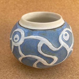 May include: A small, round ceramic pot with a blue glaze and white abstract designs. The pot has a white rim and a slightly textured surface.