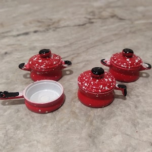 May include: A collection of miniature red and white polka-dotted toy pots and pans. The set includes three lidded pots and one frying pan with a handle. The pots have black handles and knobs. The interior of the frying pan is white.